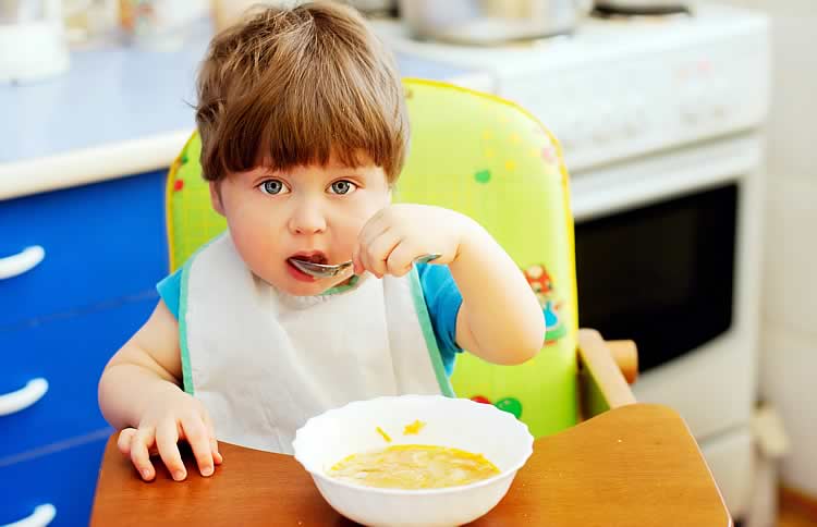 Годовалый ребенок сидит и ест суп ложкой